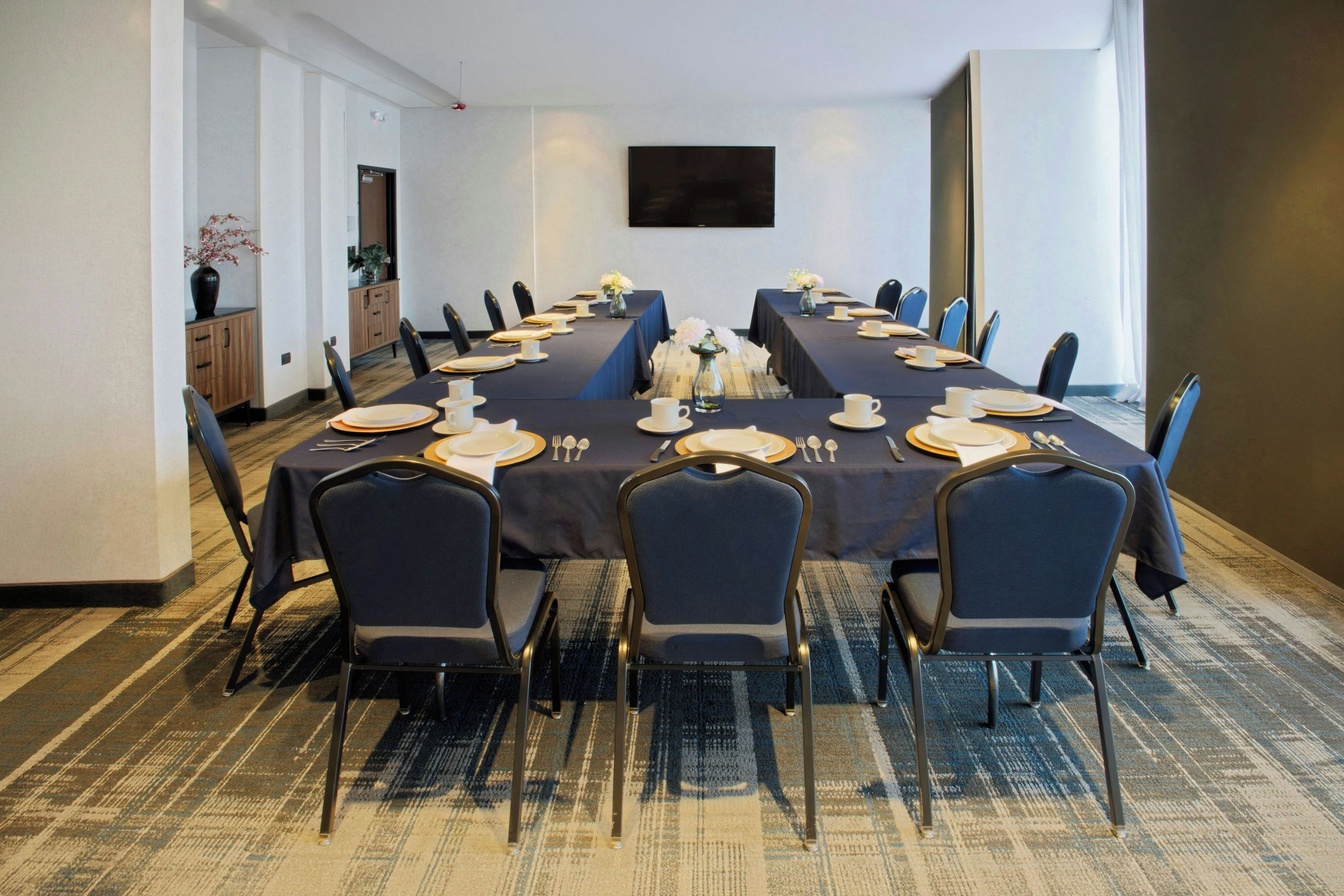 Un salón de conferencias con una larga mesa dispuesta para una reunión, equipado con sillas, platos, vasos y un jarrón con flores.