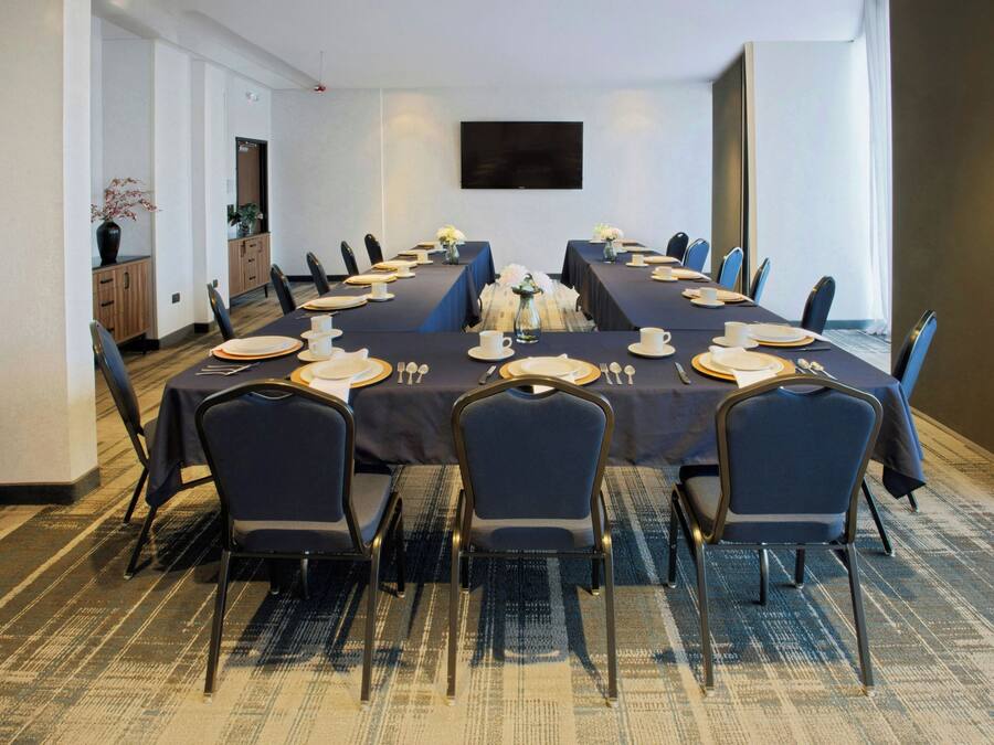 Un salón de conferencias con una larga mesa dispuesta para una reunión, equipado con sillas, platos, vasos y un jarrón con flores.