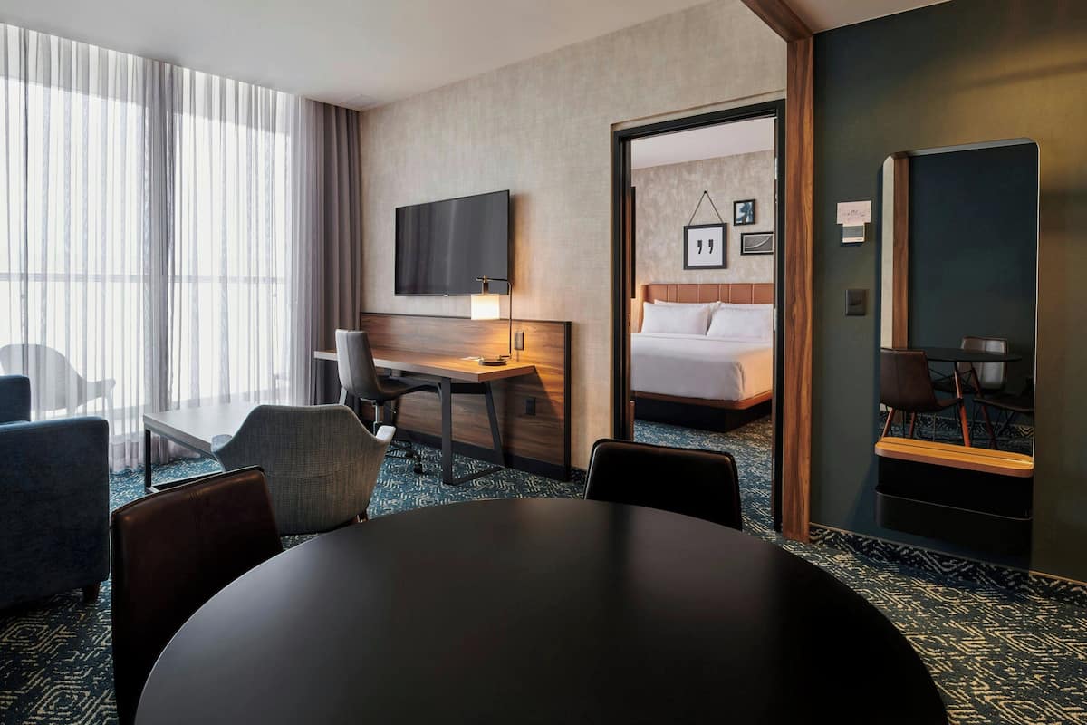A hotel room with a large bed, a desk with a chair, a TV mounted on the wall, and a dining table.