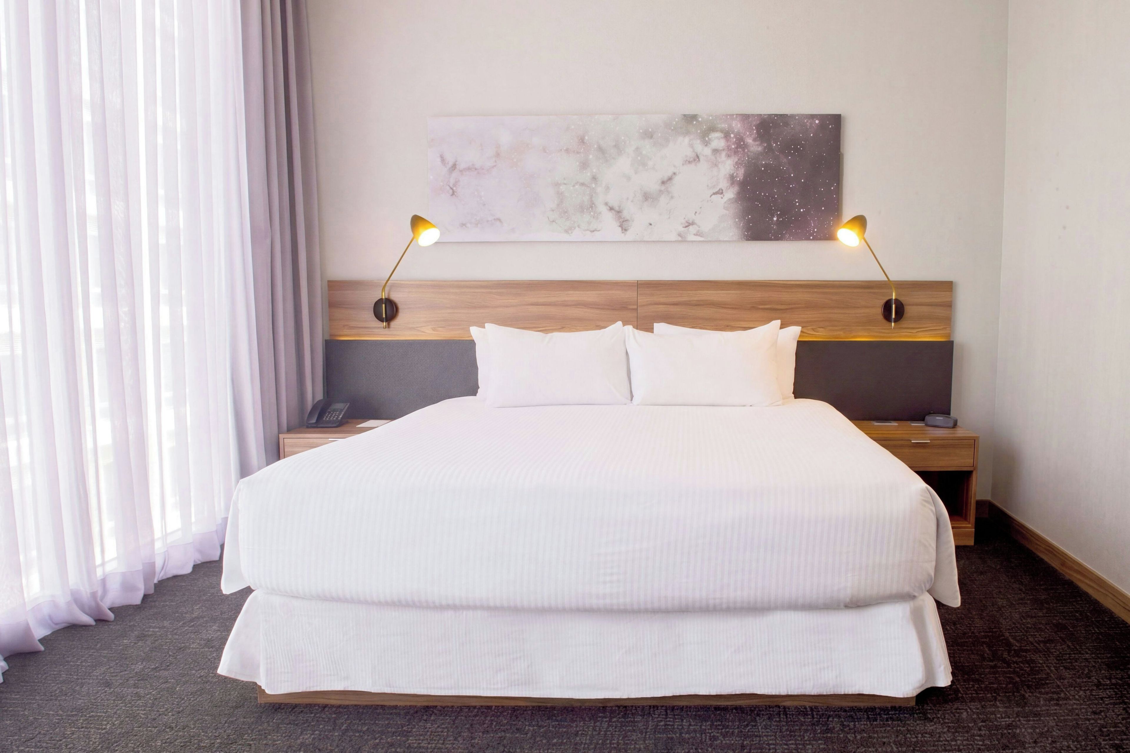 A hotel room with a large bed, two bedside lamps, a wooden headboard, a nightstand with a phone, and a painting above the bed.