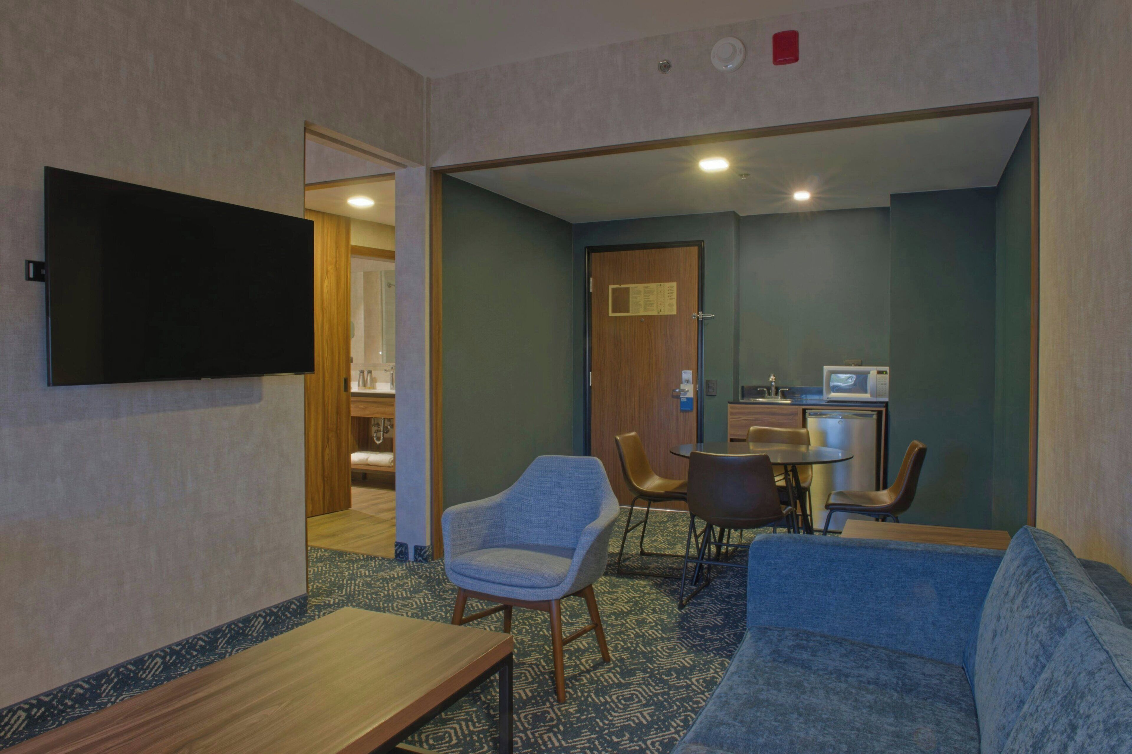 A hotel room with a flat-screen TV mounted on the wall, a sofa, a small table, and a dining area with chairs and a small kitchen.