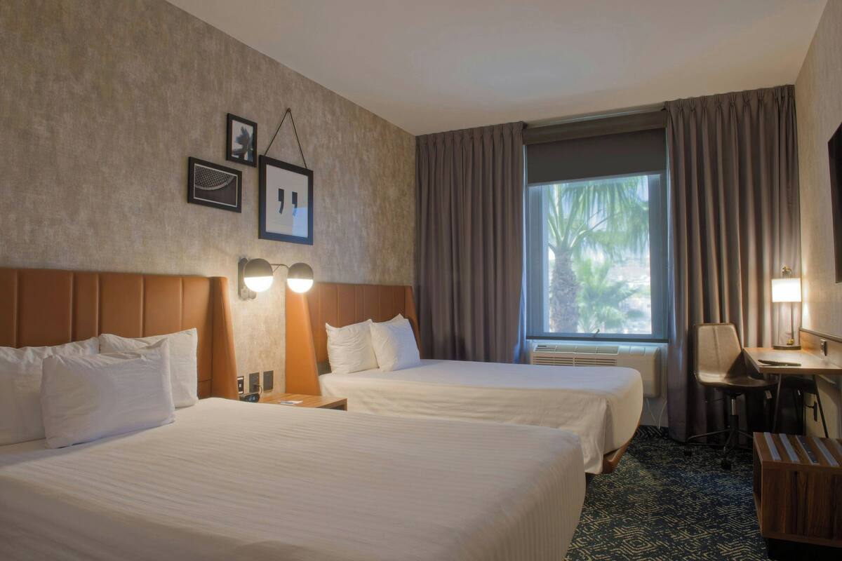 A hotel room with two beds, a desk, a chair, a lamp, and a window with curtains.