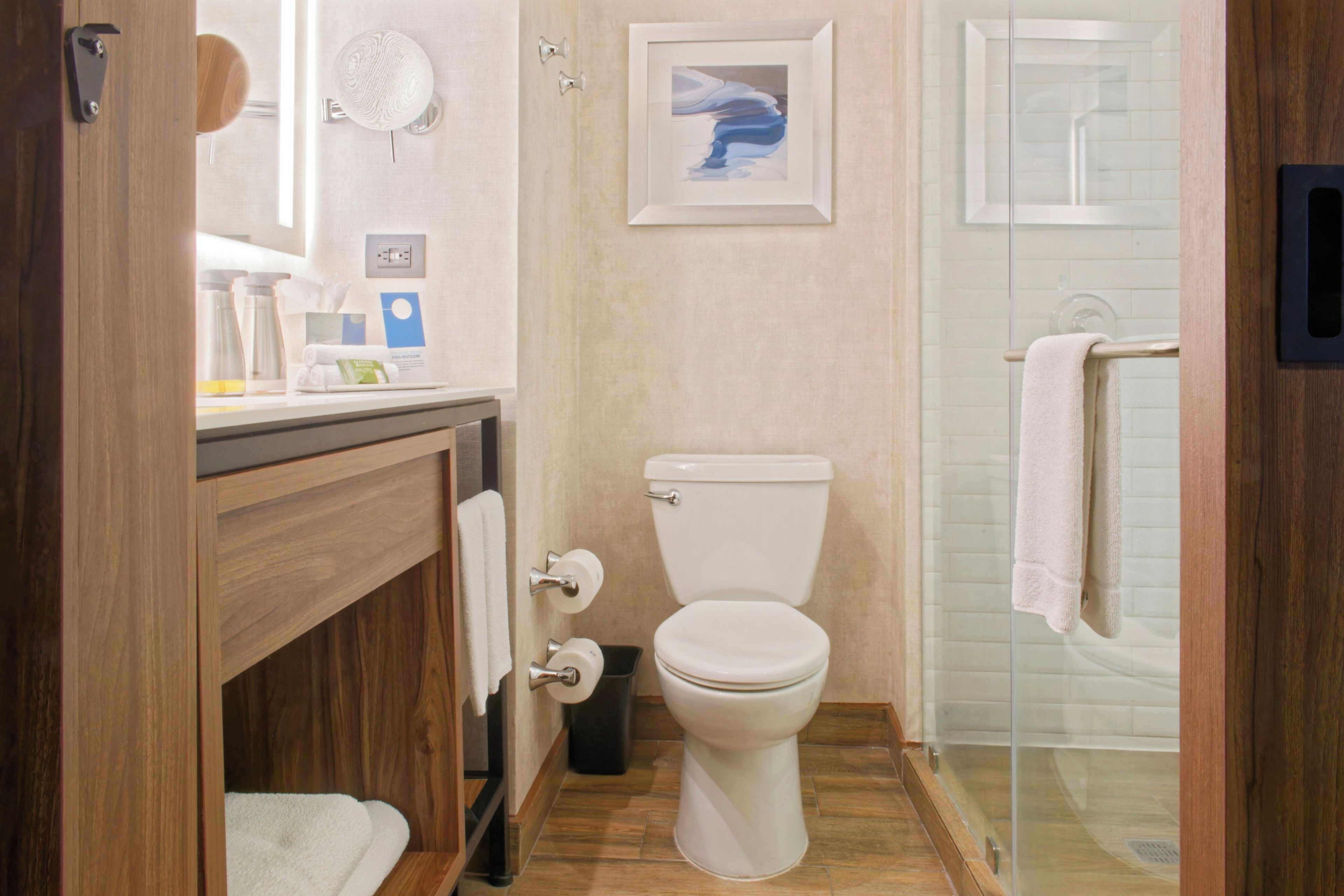 A modern bathroom with a wooden vanity, a toilet, a glass shower enclosure, and a framed artwork on the wall.