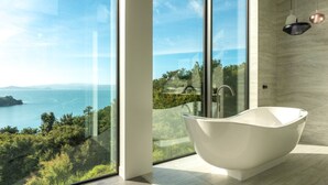 Un baño moderno con una bañera independiente, ventanas amplias que ofrecen vistas al mar y al verdor, y una decoración minimalista.