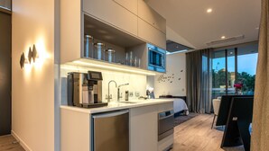 Una cocina moderna con microondas integrado, cafetera y un refrigerador pequeño. Hay un comedor con mesa y sillas, y un ventanal con vistas al exterior.