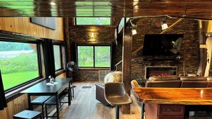 A cozy, rustic interior with a stone fireplace, leather seating, and large windows overlooking a green landscape.