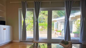 Ein Zimmer mit Glastisch, weißem Schrank und einer Schiebetür aus Glas, die zu einem Außenbereich mit Pflanzen und einem Haus führt.