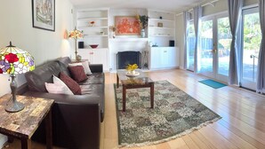 Ein Wohnzimmer mit Sofa, Couchtisch und Kamin. Regale mit Dekoration und Blick ins Freie.