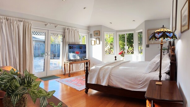 Ein Schlafzimmer mit einem großen Bett, einem Fernseher auf einem Holztisch und Blick ins Freie durch große Fenster.