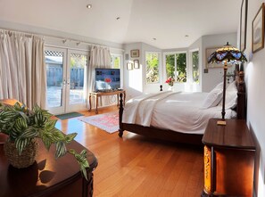 A bedroom with a large bed, a TV on a wooden table, and a view of the outdoors through large windows.