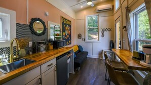 A compact kitchen with a wooden countertop, a sink, a microwave, and a small refrigerator. There is a ceiling fan, a window with curtains, and a wall-mounted air conditioner. - The Florio, Tiny Home, River, Hiking, and Rafting! (Sylva)