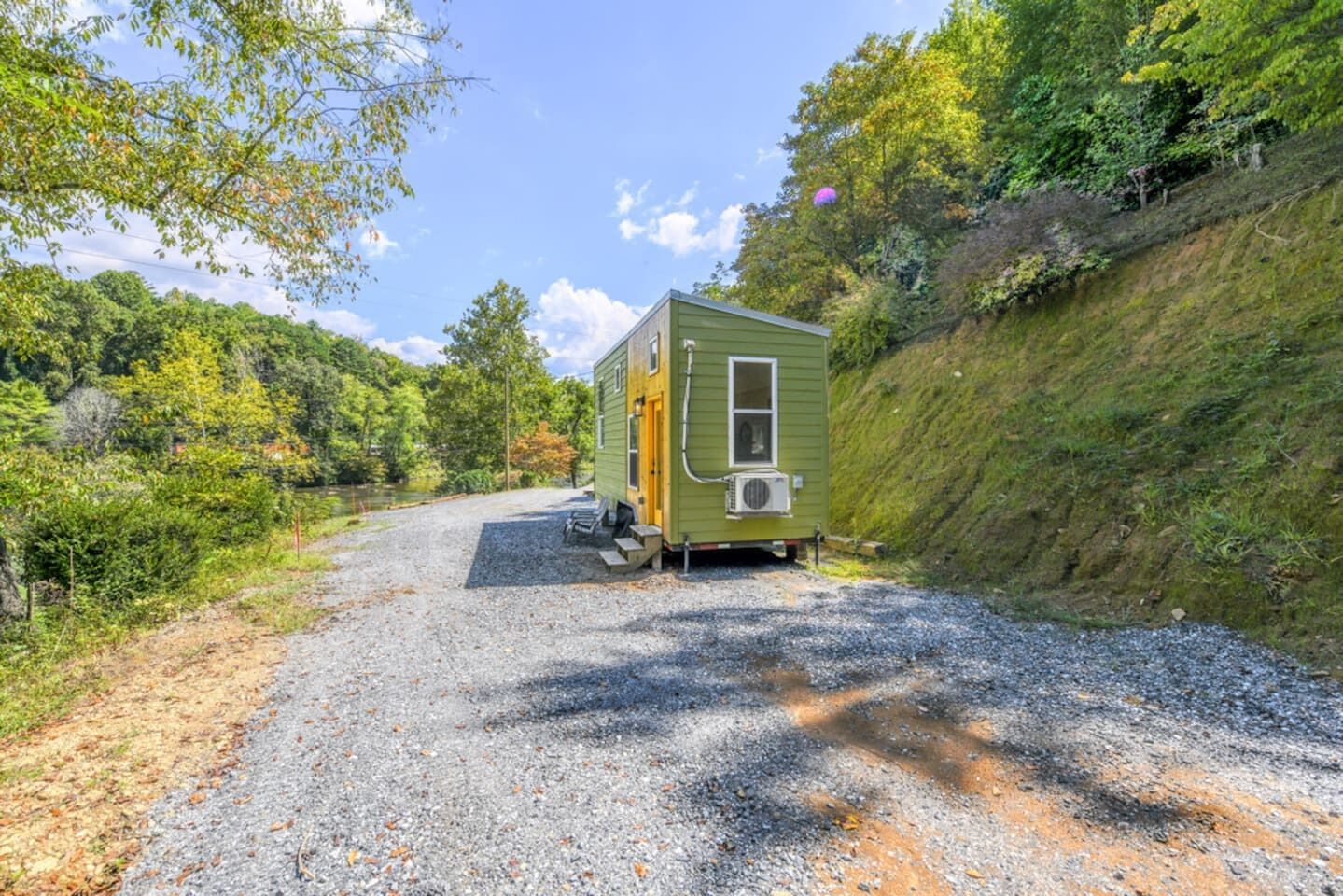 A small, single-story cabin with a front porch and a small attached structure, surrounded by a gravel driveway and lush greenery.