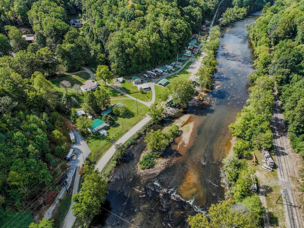 An aerial view of a riverside property with a road, parked vehicles, and a small building.