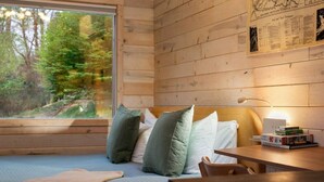 A cabin room with a bed, pillows, a table with books, a lamp, and a window with a view of trees.