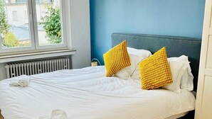 Egy gondosan megterített ágy fehér ágyneművel és két feltekert törölközővel, egy kék színű fal, és egy a mennyezetről lógó lámpatest.