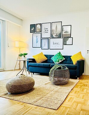 A living room with a blue sofa, two round poufs, a woven coffee table, and a wall decorated with framed pictures and a lamp. - Elegant one bedroom w private terrace (Luxembourg City)