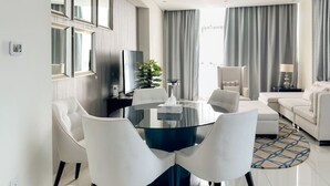 Moderni hotellihuone, jossa on pyöreä ruokapöytä, valkoiset tuolit, sohva ja televisio.