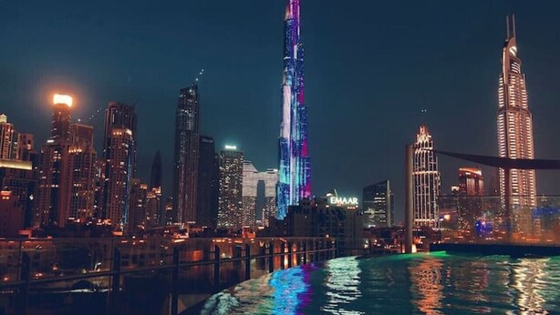 A nighttime cityscape with illuminated skyscrapers, including a prominent tower with a spire, reflecting on a body of water. - Panoramic Burj Khalifa View Luxury Penthouse 1BR (Dubai)