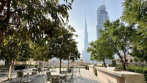 An outdoor seating area with tables and chairs, surrounded by trees, overlooking a cityscape with a prominent skyscraper. - Stunning Burj View Sky High Luxury 2BR (Dubai)