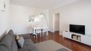 Una sala moderna con un sofá gris, una mesa de comedor con sillas, un televisor de pantalla plana y un mueble blanco para entretenimiento.