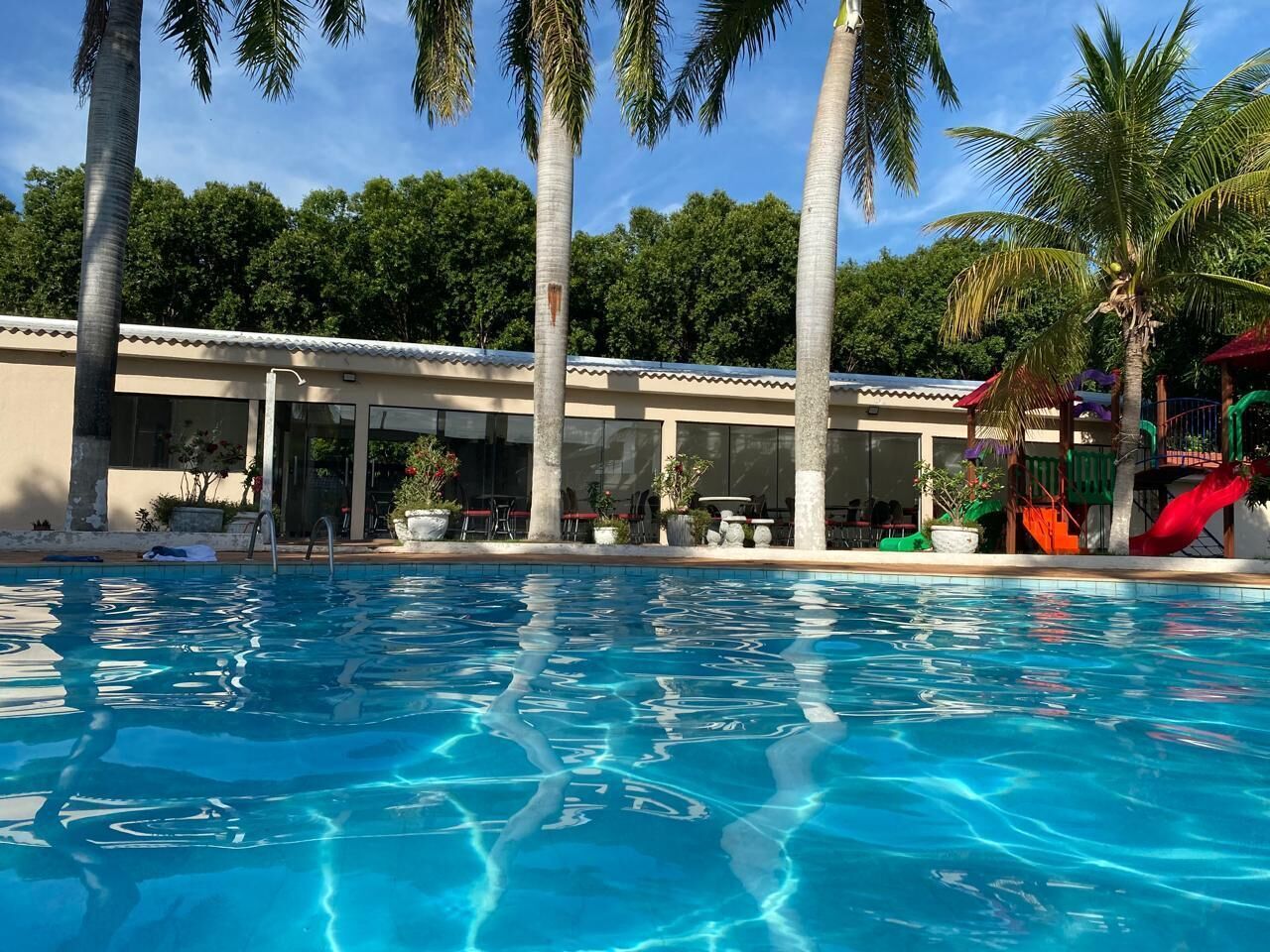 A swimming pool with clear blue water, surrounded by palm trees and a building with outdoor seating.