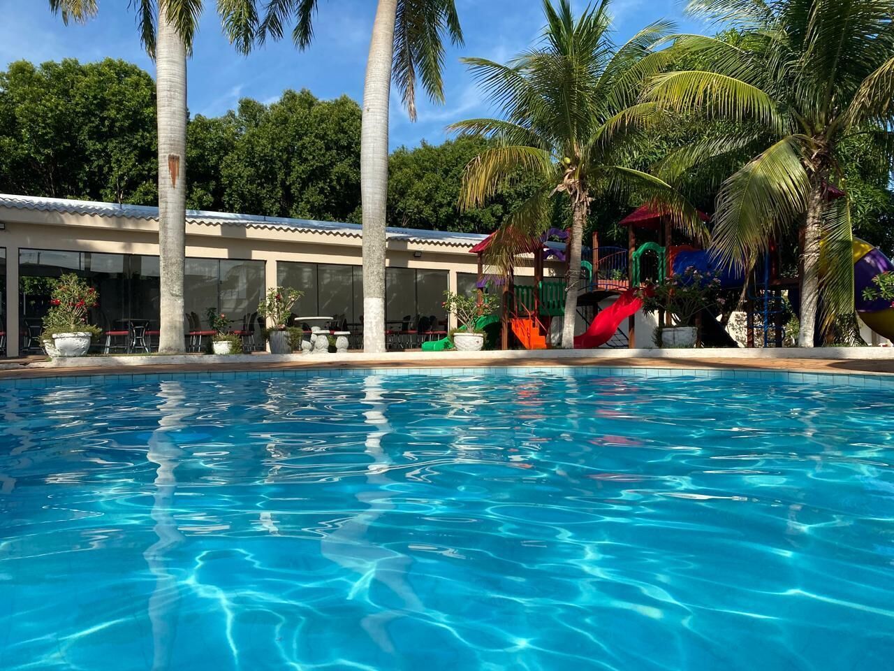 Una piscina con agua cristalina azul, rodeada de palmeras y un edificio con techo de tejas.