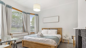 Une chambre à coucher avec un lit en bois, un bureau en verre, une table de chevet blanche, une cheminée en briques noires et une fenêtre avec des rideaux.