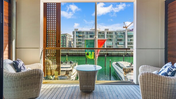 Un balcon doté de meubles en osier, d’une balustrade en verre et offrant une vue sur les bateaux et les bâtiments.