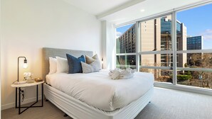 A modern hotel room with a large bed, a bedside table with a lamp, and a view of a cityscape through large windows. - Chic Central Apt with Gym and Pool by Zodiak Stays (Auckland)