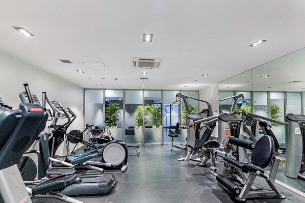 A modern gym with various exercise machines, a large mirror, and a view of the outside through glass doors.