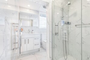 A modern bathroom with a glass shower enclosure, white marble tiles, and a white vanity with a mirror. - Ellerslie 3BR House | Fresh Reno | Zodiak Stays (Auckland)