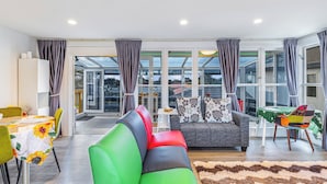 A modern living room with a sofa, a dining table with chairs, and a view through large windows. - Ellerslie 3BR House | Fresh Reno | Zodiak Stays (Auckland)
