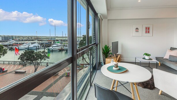 A modern room with a large window overlooking a marina, a round table with chairs, a sofa, and a small side table with a plant. - Viaduct Harbour Views w/ Free Parking-Zodiak Stays (Auckland)