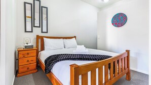 A neatly made bed with white linens, a wooden headboard, and a black knitted blanket. There is a wooden nightstand with drawers and a lamp beside the bed. Three framed pictures are hung on the wall above the bed. - 1BR Grey Lynn Haven+Parking, Balcony Zodiak Stays (Auckland)