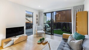 A modern living room with a flat-screen TV, a grey sofa, a wooden coffee table, and a sliding glass door leading to a balcony with a view of a house and a garden.