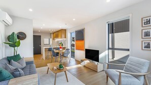 A modern living room with a sofa, a coffee table, and a TV. There is a dining area with chairs and a table in the background.