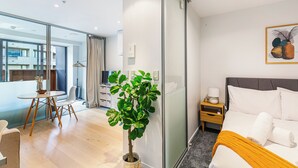A modern hotel room with a bed, bedside table, and a potted plant. There is a dining area with a table and chairs, and a view of the city through large windows. - Sleek central stay with balcony by Zodiak Stays (Auckland)