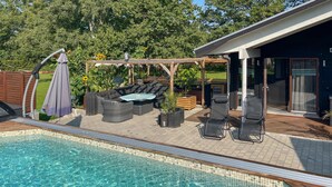 Una piscina con un sillón de sol, una zona de estar cubierta y un patio con sillas y una mesa.
