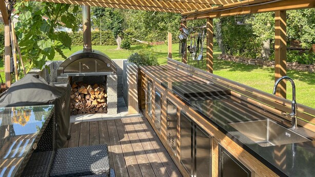 Un área de cocina al aire libre techada, con una parrilla, cubierta de madera y un pérgola.