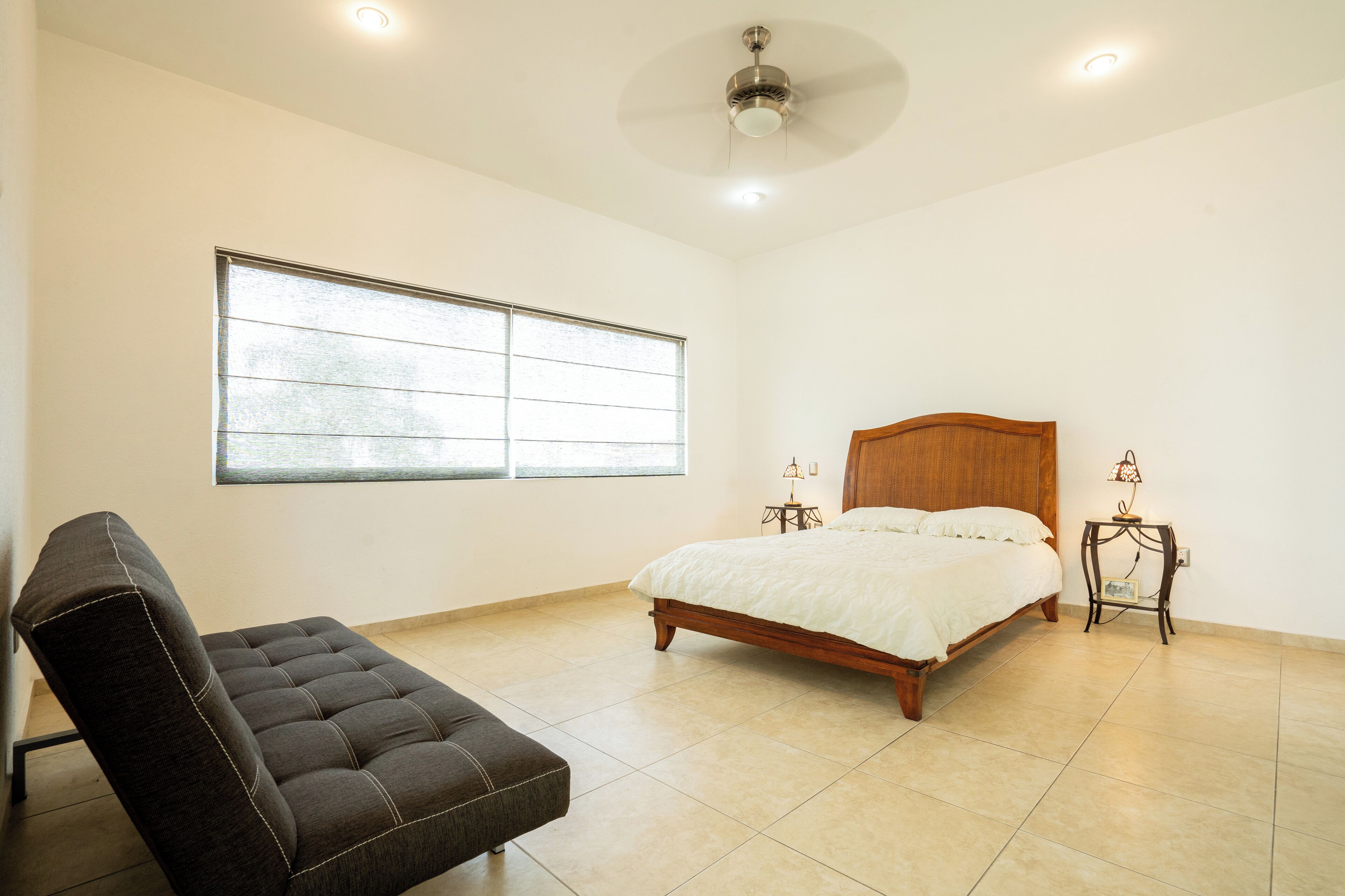 Un dormitorio con cama, una silla, un ventanal y un ventilador de techo.