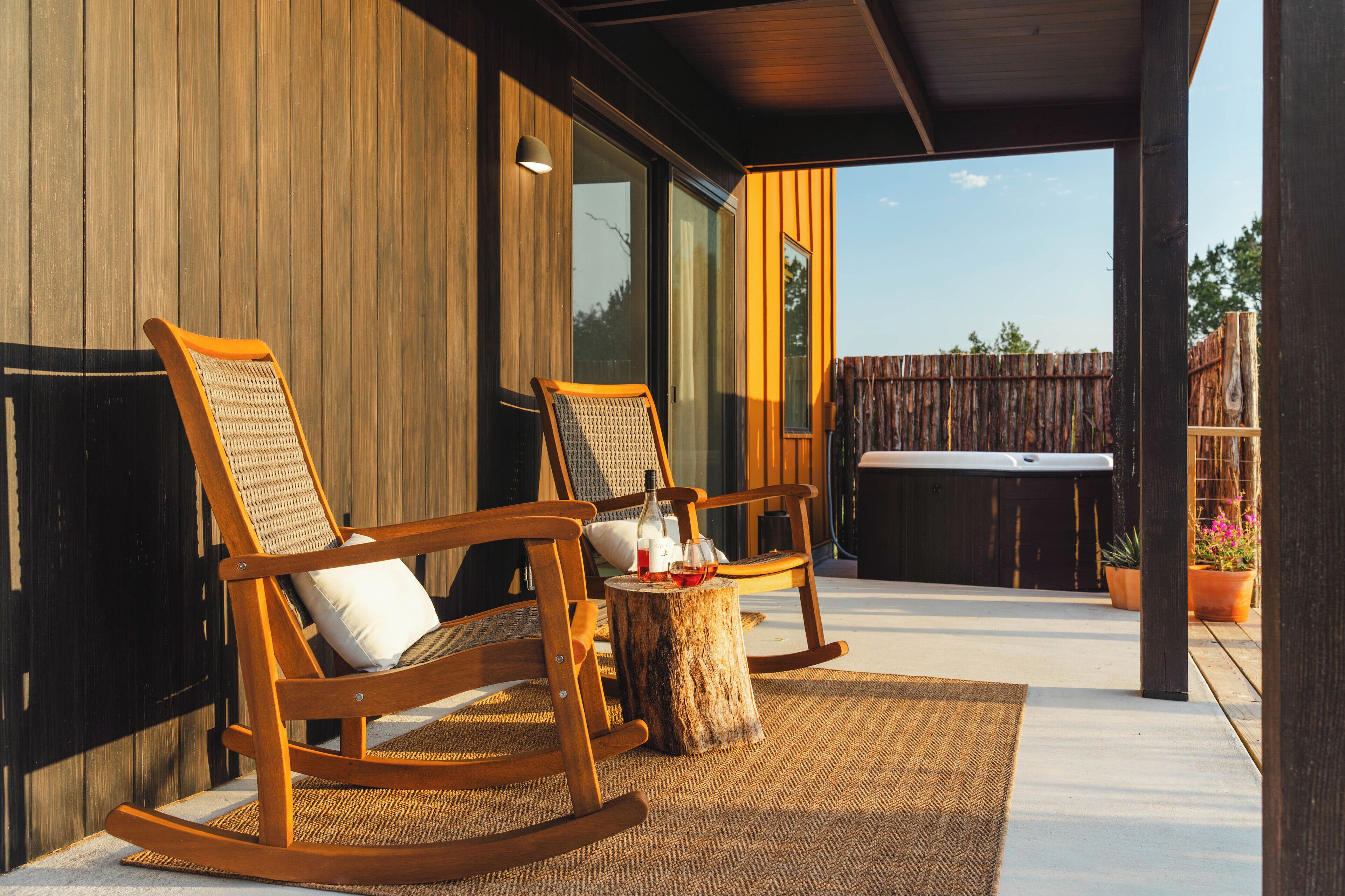 A wooden rocking chair and a second chair with cushions, a small wooden table with a bottle and glasses, and a view of a wooden fence and trees in the background.