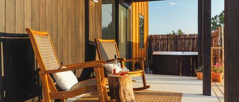 A wooden rocking chair and a second chair with cushions, a small wooden table with a bottle and glasses, and a view of a wooden fence and trees in the background.