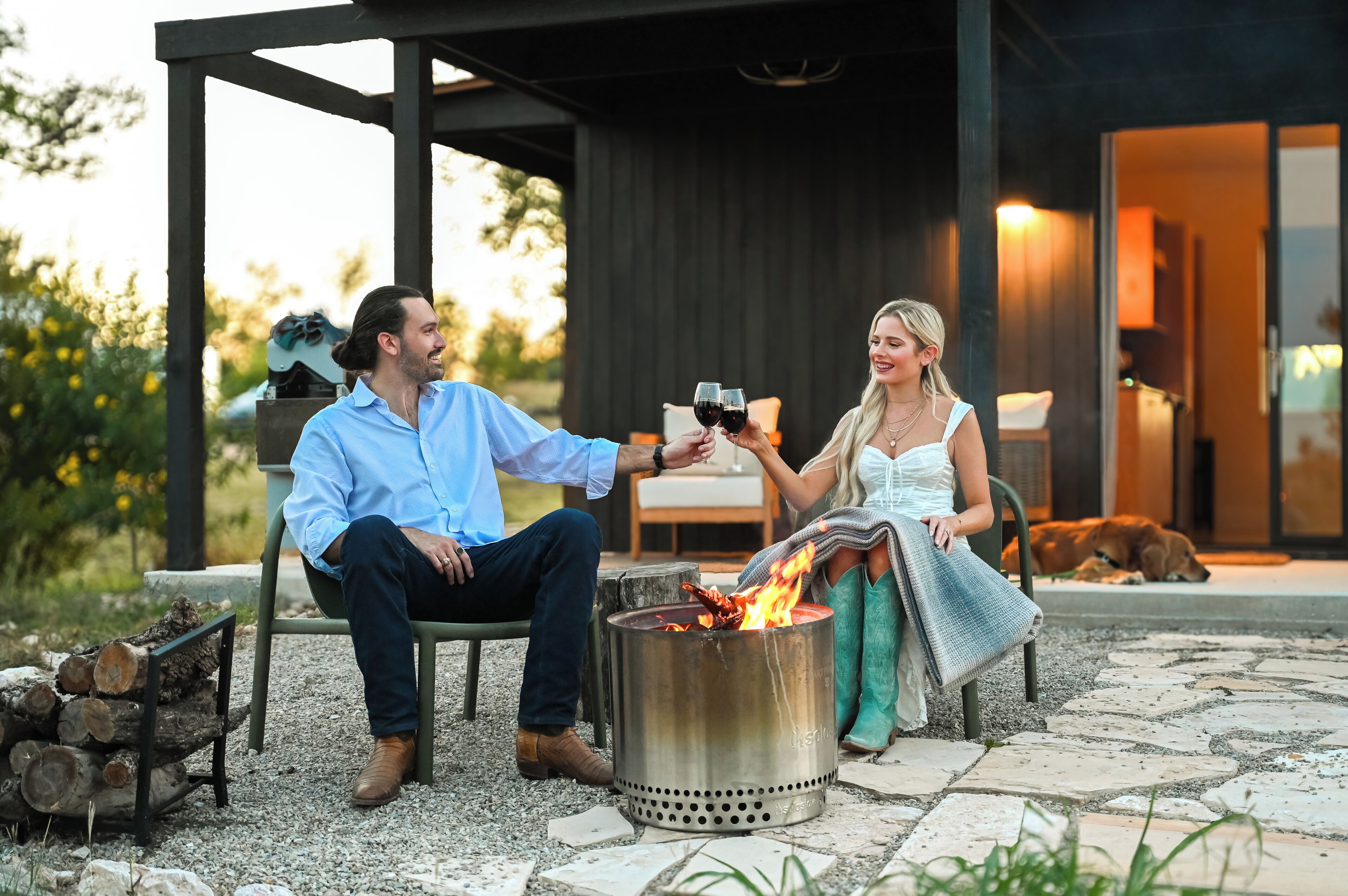 A couple seated by a fire pit in an outdoor setting with a modern building in the background.