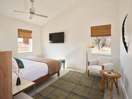 A bedroom with a bed, bedside tables, a chair, a floor lamp, a TV, and windows with blinds.
