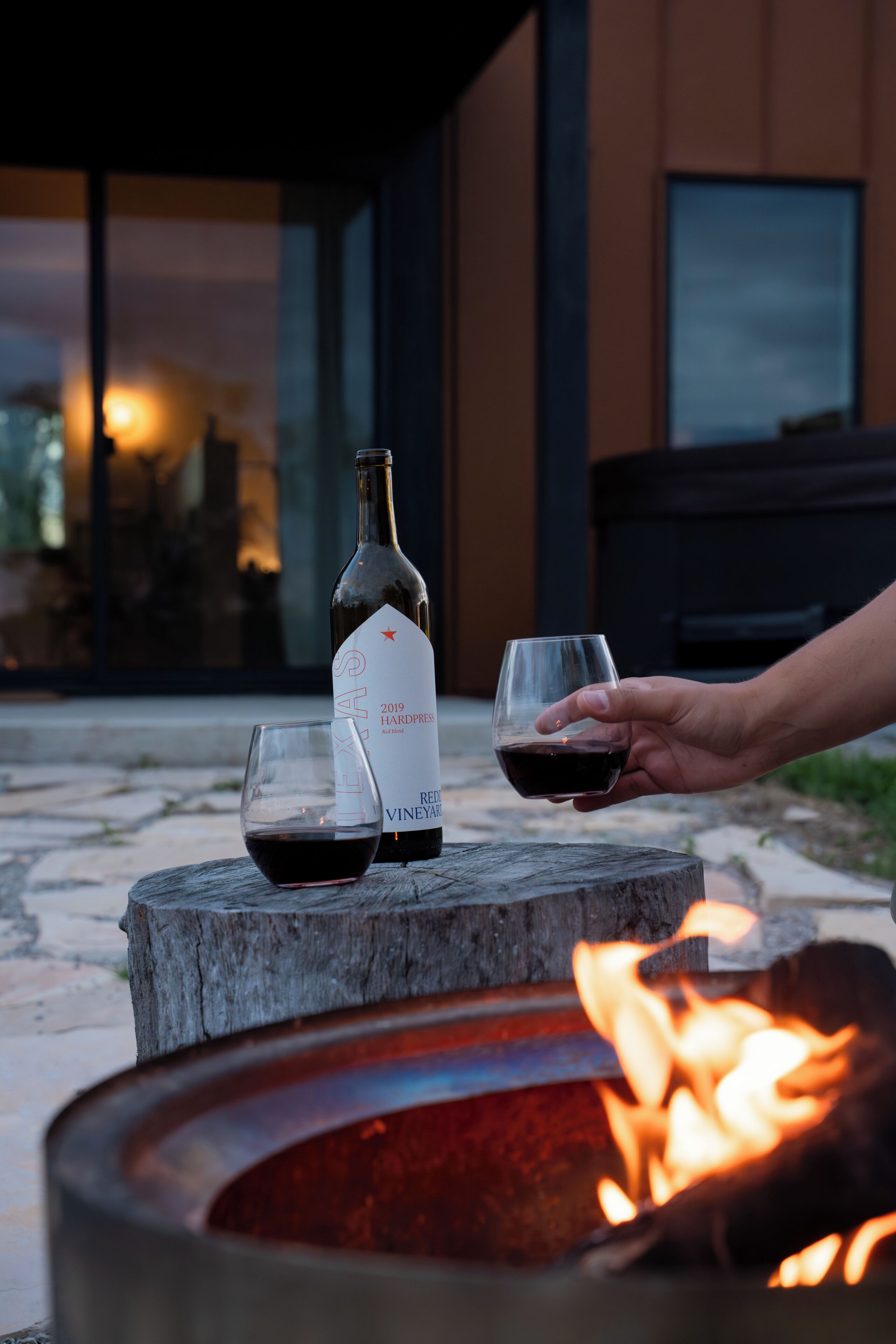 A bottle of wine and two glasses on a wooden stump with a fire pit in the background.
