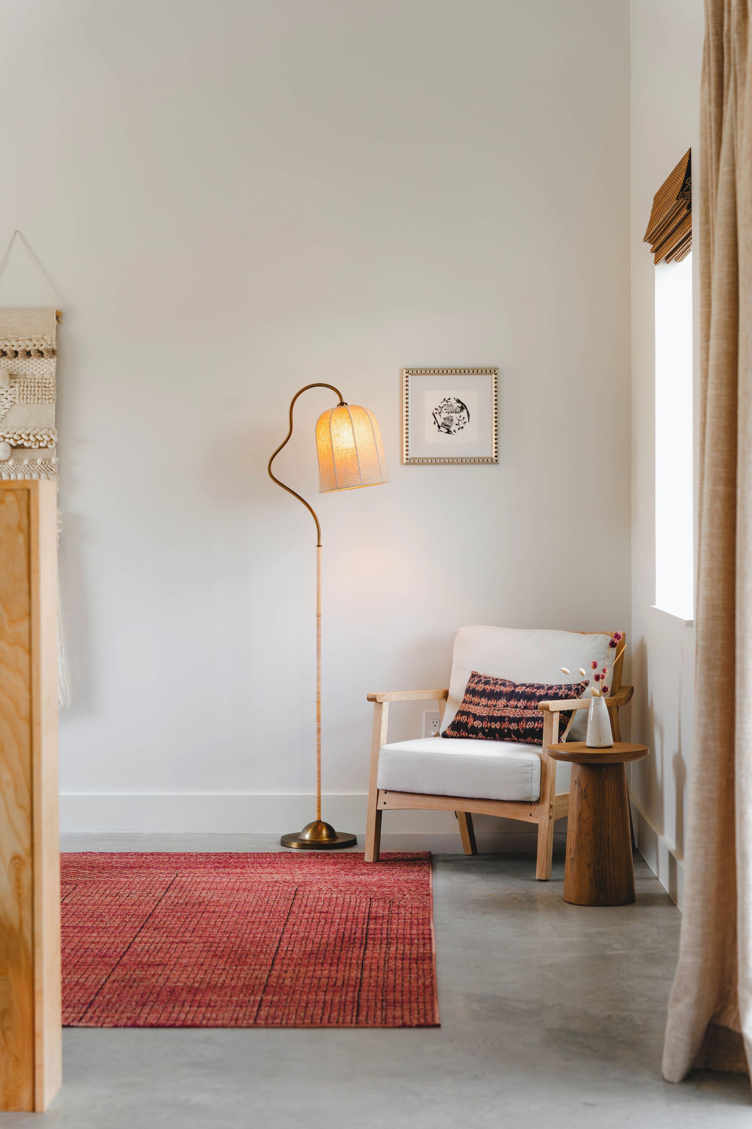 A room with a wooden floor, a red rug, a wooden chair with a cushion, a floor lamp, and a framed picture on the wall.