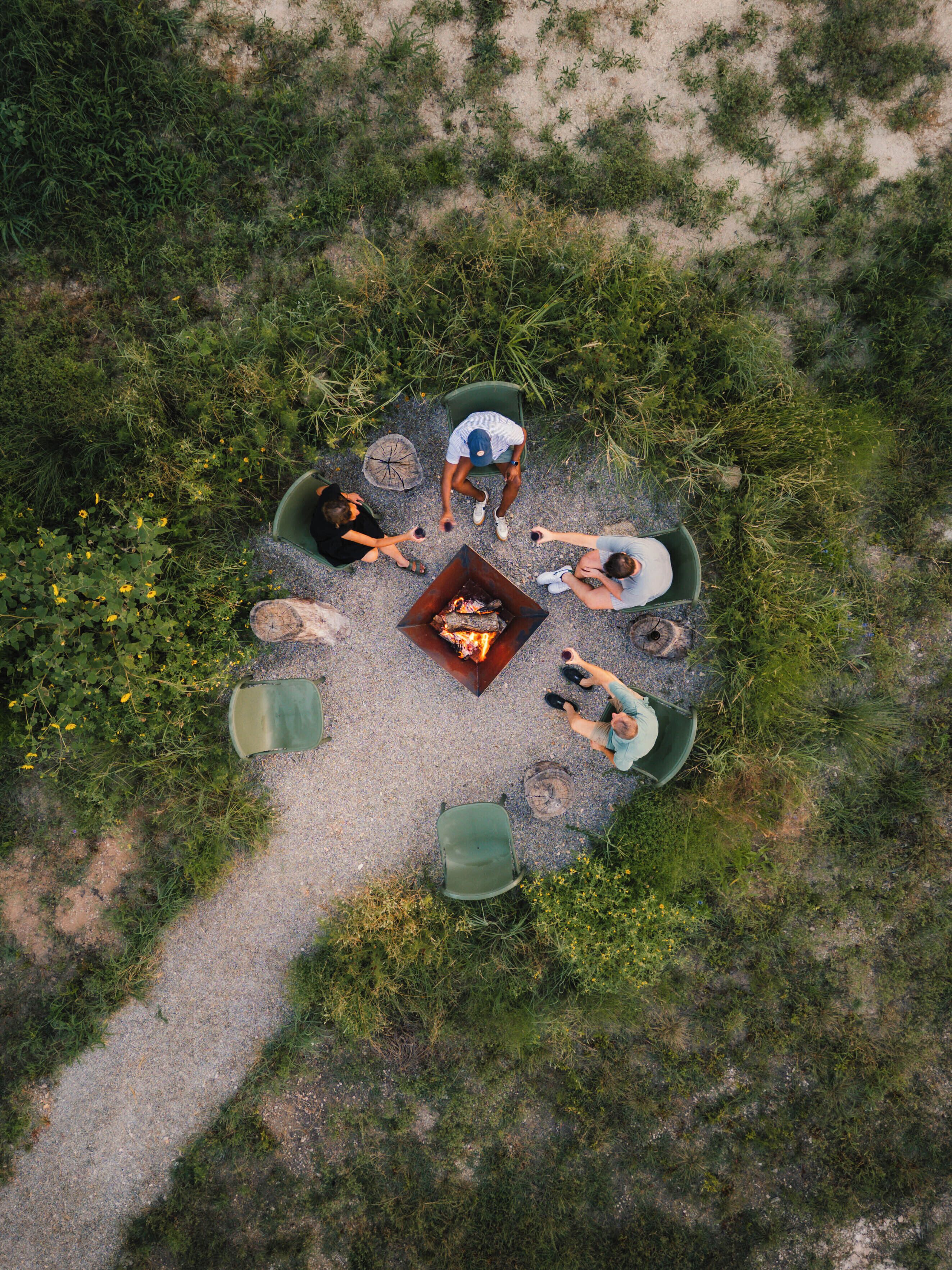 Vista aérea de uma área de estar circular ao ar livre, com uma fogueira central, rodeada por vegetação e cascalho.