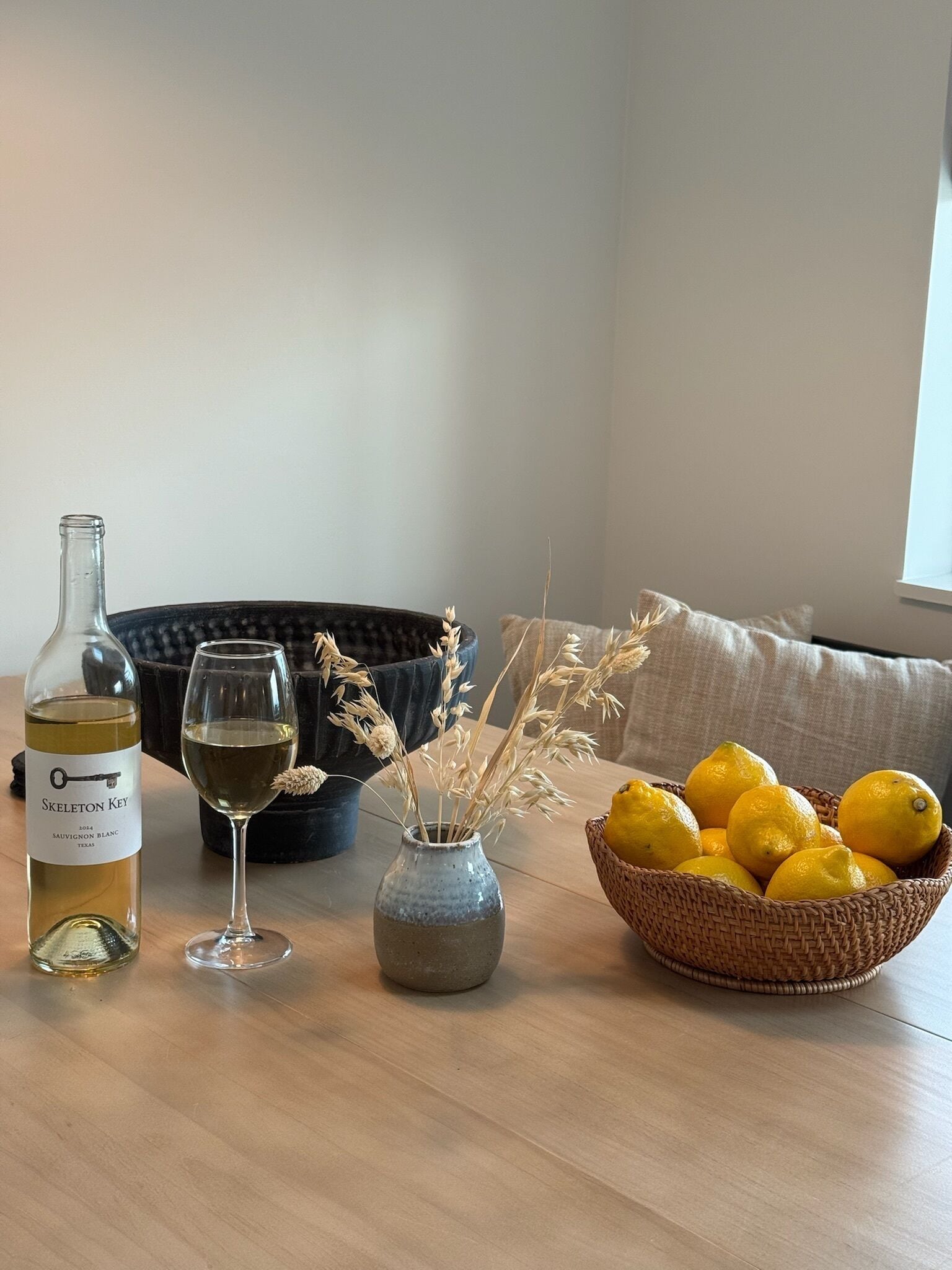 A bottle of wine, a glass, a vase with dried plants, and a basket of lemons on a wooden table.