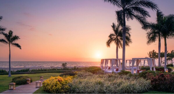 Vista al atardecer con playa, palmeras y zonas de estar al aire libre.