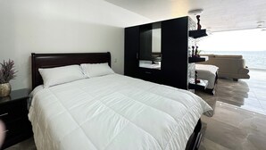 Una habitación de hotel moderna con una cama grande, un área de descanso con vistas al mar y una pequeña mesa con objetos decorativos.